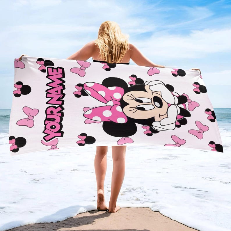 May include: A rectangular beach towel featuring a white background with a repeating pattern of pink bows and black Minnie Mouse silhouettes. The towel displays a large graphic of Minnie Mouse and the text YOUR NAME in bold pink letters.