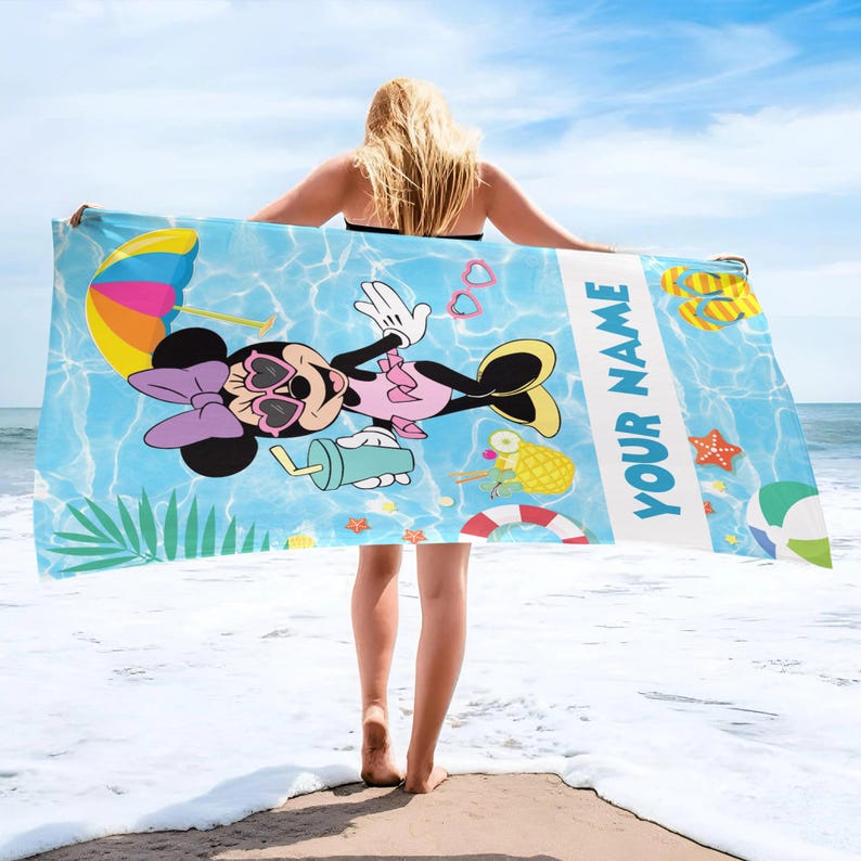 Op de afbeelding: Een strandlaken met Minnie Mouse met zonnebril, een roze jurk en een paarse strik, tegen een blauwe waterachtergrond met strandillustraties. De tekst "YOUR NAME" staat op de handdoek.