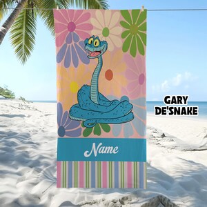 May include: A beach towel featuring a cartoon snake character in shades of blue, set against a backdrop of large, colorful flowers. The towel has a blue stripe with the word "Name" and a striped border. The text "GARY DE'SNAKE" is also visible.