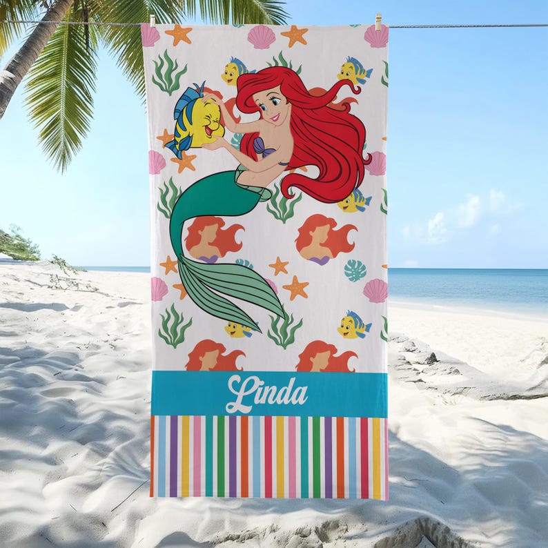 K&ouml;nnte beinhalten: Wei&szlig;es Strandtuch mit einer Cartoon-Meerjungfrau mit roten Haaren und einem gr&uuml;nen Schwanz, die einen gelben Fisch h&auml;lt. Das Handtuch hat einen blauen Streifen mit dem Namen "Linda" und einen bunten gestreiften Rand. Dekorative Elemente sind Seesterne, Muscheln und Seetang.