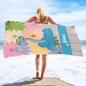 May include: A beach towel featuring a cartoon blue snake with a flower design background. The towel has a blue stripe with the word "Name" in white script and colorful stripes. The towel is held up by a person on a beach.