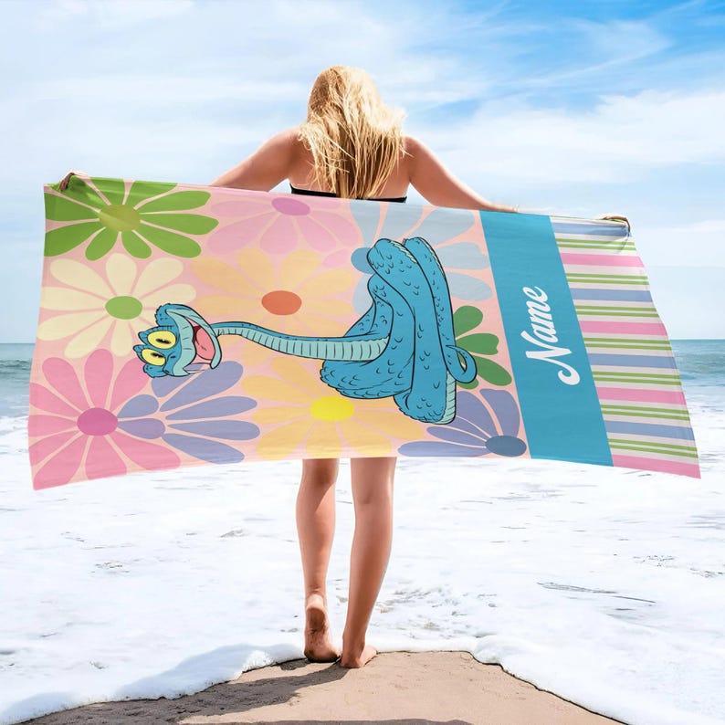 May include: A beach towel featuring a cartoon blue snake with a flower design background. The towel has a blue stripe with the word "Name" in white script and colorful stripes. The towel is held up by a person on a beach.