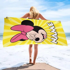 Puede incluir: Una toalla de playa rectangular con un fondo de patr&oacute;n de rayos de sol amarillo brillante. La toalla muestra un gr&aacute;fico grande de Minnie Mouse con un lazo rosa y la palabra NAME impresa en letras may&uacute;sculas blancas en el lado derecho.