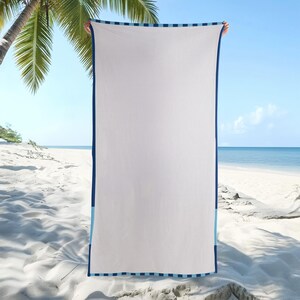 May include: A white beach towel with a navy blue border and light blue stripes. The towel is rectangular and held up against a beach backdrop with palm trees and ocean views.