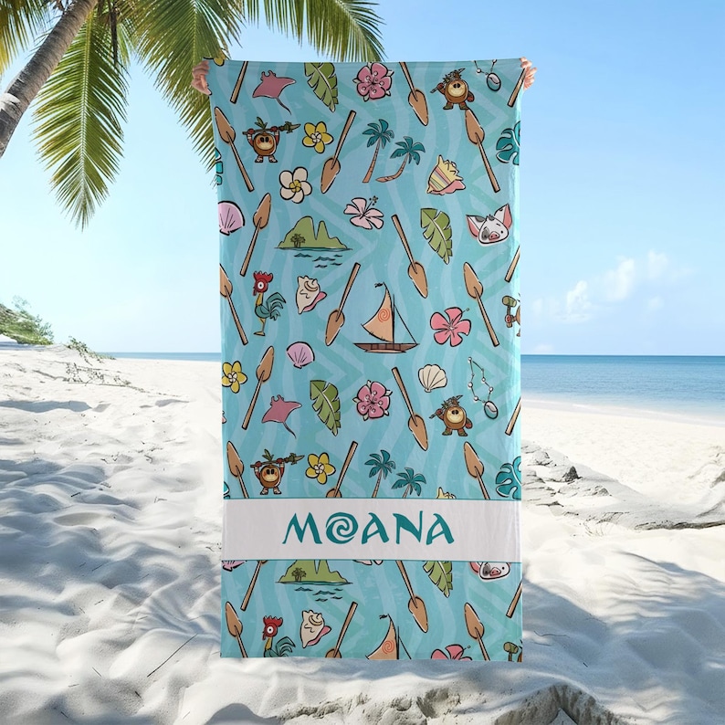 May include: A light blue beach towel with a repeating pattern of cartoon characters, flowers, and boats. The word "MOANA" is printed in white on a white band at the bottom of the towel. The towel is held up on a beach with palm trees and the ocean in the background.
