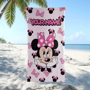 May include: A light pink rectangular beach towel featuring a large graphic of Minnie Mouse wearing a pink and white polka dot bow. The towel is patterned with small black mouse ears and pink bows, with the text YOUR NAME printed in black.
