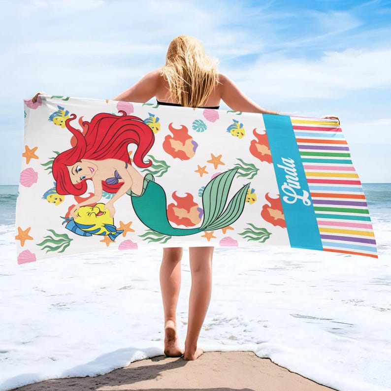 K&ouml;nnte beinhalten: Ein wei&szlig;es Strandtuch mit einer Cartoon-Meerjungfrau mit roten Haaren und einem gr&uuml;nen Schwanz, mit einem gelben Fisch. Das Handtuch hat einen blauen Streifen mit dem Namen "Linda" und einen gestreiften Rand in verschiedenen Farben. Das Handtuch wird von einer Person am Strand gehalten.