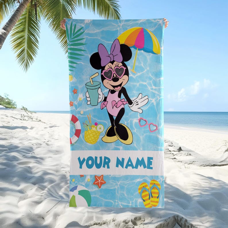 Op de afbeelding: Strandlaken met Minnie Mouse in badpak, zonnebril en strik. De achtergrond is blauw met strandthema graphics en de tekst "YOUR NAME". Inclusief een kleurrijke parasol en drankje.