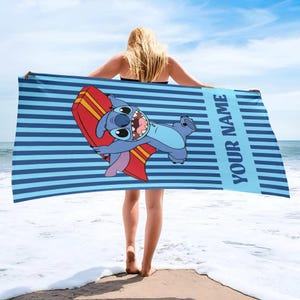 May include: Blue beach towel with horizontal stripes and the text "YOUR NAME" in a vertical blue stripe. The towel features a cartoon character holding a red and yellow surfboard. The character is blue with large eyes and a wide grin.