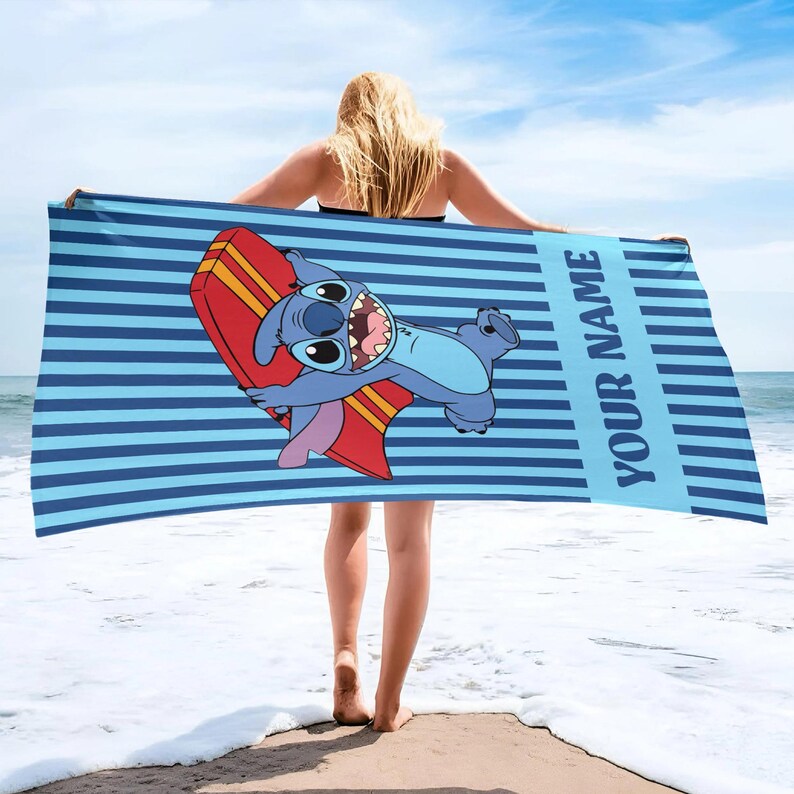 May include: Blue beach towel with horizontal stripes and the text "YOUR NAME" in a vertical blue stripe. The towel features a cartoon character holding a red and yellow surfboard. The character is blue with large eyes and a wide grin.