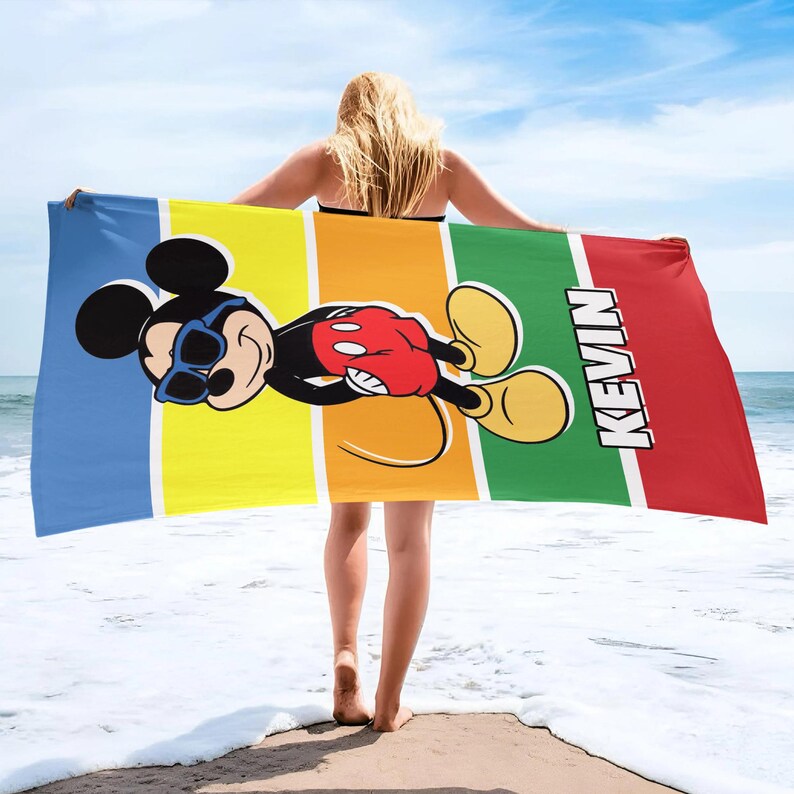 May include: A vibrant beach towel with a cartoon Mickey Mouse design, wearing sunglasses, set against a backdrop of blue, yellow, orange, green, and red stripes. The name "KEVIN" is printed vertically in white. The towel is held up by a person on a beach.