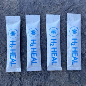 以下が含まれることがあります： 青い文字と青いグラフィックが入った白いパケットが4つ。パケットには青色で「H2 HEAL」と書かれており、その上に円形のグラフィックがあります。パケットは上部が密封されており、灰色の表面に置かれています。