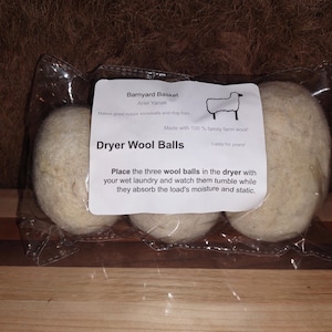 May include: Three off-white dryer wool balls in a clear plastic package. The label reads "Dryer Wool Balls" and includes instructions. The package is on a wooden surface with a brown background.