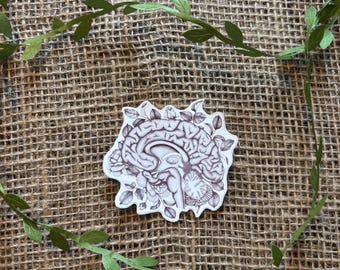 Op de afbeelding: Een sticker met een gedetailleerde illustratie van een menselijk brein, omringd door bloemmotieven. De sticker is in grijstinten en wit, geplaatst tegen een bruine achtergrond met groene bladguirlande.
