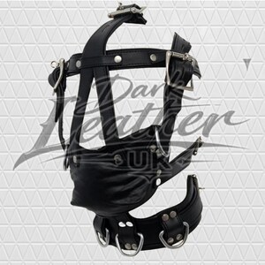 May include: A black leather head harness featuring a central mouth covering, adjustable straps with silver-toned metal buckles, rivets, and D-rings. The structure includes a neck collar section and a vertical strap assembly with Dark Leather UK text.