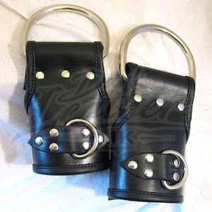 May include: Pair of black leather wrist restraints with silver-colored metal accents. Each restraint features a large, circular ring and a buckle closure. The leather is detailed with rivets.