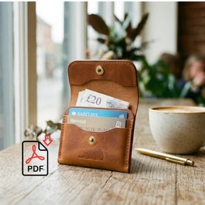 May include: A brown leather wallet with a snap closure, holding a twenty-pound note and several credit cards. The wallet is open, revealing its interior. A gold pen and a cup of coffee are on the wooden table.