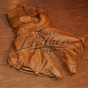 May include: A tan leather restraint suit featuring multiple adjustable straps with metal buckles, D-rings, and a high collar with a central zipper. The garment includes a lace-up front section and is branded with the text Dark Leather UK.