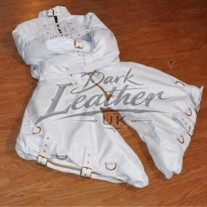 May include: A white straightjacket featuring long sleeves, multiple adjustable straps with gold-toned metal buckles, and reinforced eyelets. The garment includes a high collar with a buckle closure and a central lacing system on the upper chest area.