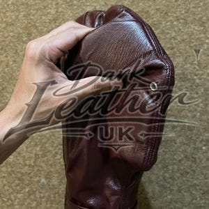 May include: A dark brown leather glove featuring a smooth, textured surface with a metallic silver eyelet on the side and a strap at the wrist secured by a silver metal D-ring and two rivets. The leather has a visible grain and sheen.