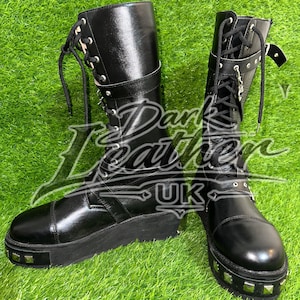 May include: A pair of black mid-calf boots featuring a lace-up front, a thick platform sole with silver-toned square studs along the perimeter, and a buckled strap near the top. The boots are marked with the text Dark Leather UK across the center.