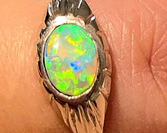 May include: A silver-toned ring featuring an oval-shaped opal gemstone with vibrant green and yellow flashes of colour. The metal band displays a textured, grooved design surrounding the central stone, creating a distinct metallic frame.