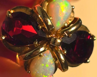 May include: A gold ring featuring a cluster of four pear-shaped gemstones arranged in a cross pattern. The design includes two deep red garnet stones and two iridescent white opals, all securely held in place by polished gold metal prongs.
