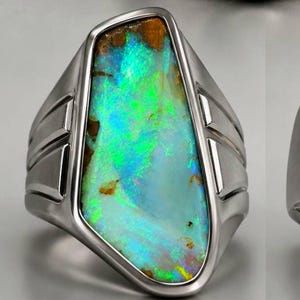 May include: A collection of five silver rings, each featuring a unique, raw, multi-colored opal gemstone. The stones display vibrant flashes of blue, green, and brown, set within polished silver bands with geometric side detailing and prongs.