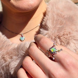 May include: A silver ring featuring a central iridescent opal surrounded by small, multi-coloured gemstones in a star-like setting, paired with a matching silver necklace pendant that also displays a single iridescent opal and a small red stone.