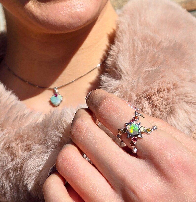 May include: A silver ring featuring a central iridescent opal surrounded by small, multi-coloured gemstones in a star-like setting, paired with a matching silver necklace pendant that also displays a single iridescent opal and a small red stone.