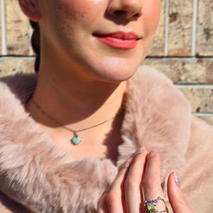 May include: A silver ring featuring a central iridescent opal stone surrounded by small colourful accent gems on a delicate band, paired with a matching necklace featuring a small heart-shaped pendant with a light blue stone and a pink accent.