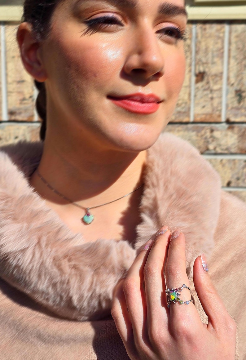 May include: A silver ring featuring a central iridescent opal stone surrounded by small colourful accent gems on a delicate band, paired with a matching necklace featuring a small heart-shaped pendant with a light blue stone and a pink accent.