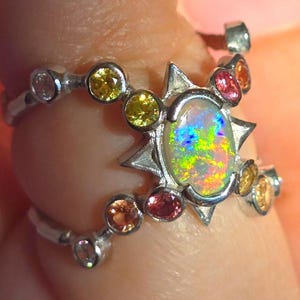 May include: A silver ring featuring a central oval opal with iridescent flashes of green, blue, and orange. The setting is accented by a starburst frame and multiple round-cut gemstones in shades of yellow, pink, and orange along the band.