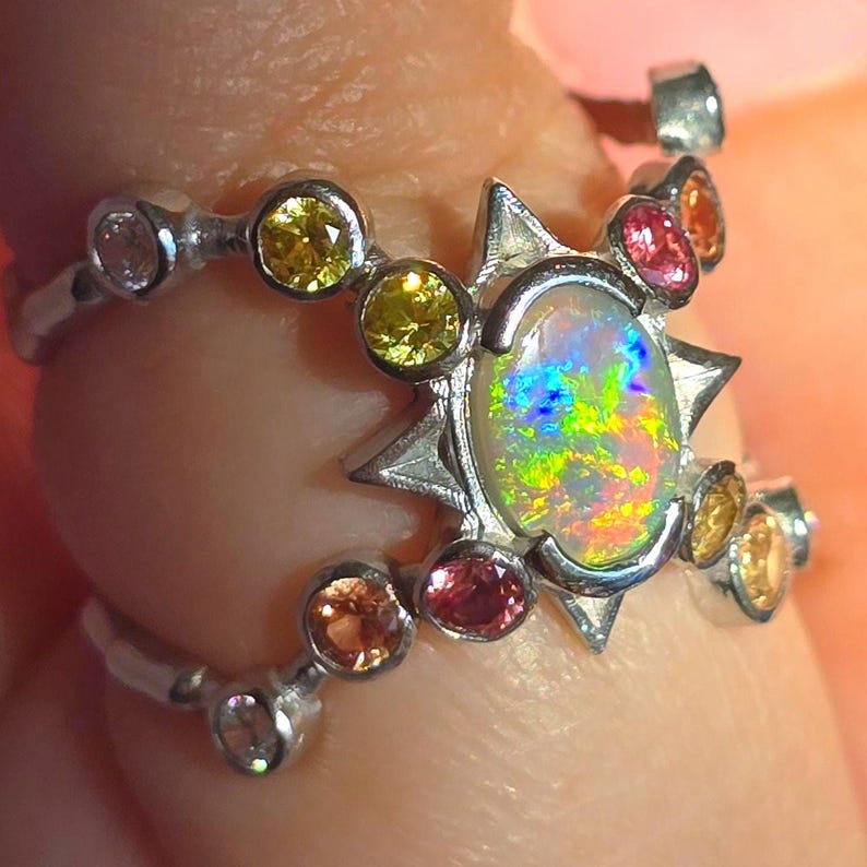 May include: A silver ring featuring a central oval opal with iridescent flashes of green, blue, and orange. The setting is accented by a starburst frame and multiple round-cut gemstones in shades of yellow, pink, and orange along the band.