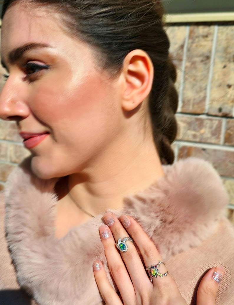 May include: Two silver rings worn on fingers. The upper ring features a single oval-shaped iridescent opal stone with a dark blue-green play of colour. The lower ring is a wider band set with multiple small, multicoloured opal stones.