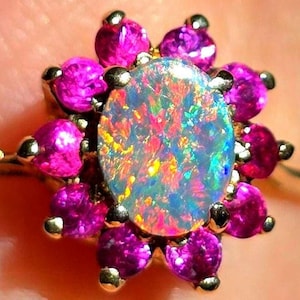 May include: A gold ring featuring an oval opal gemstone at the center, surrounded by vibrant pink gemstones. The opal displays a spectrum of colors, including blue, green, and red. The ring's design is floral, with the pink gemstones arranged around the opal.
