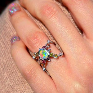 May include: A silver ring featuring a central oval opal with a vibrant play-of-colour, surrounded by a star-shaped setting. The band is accented with small, round, multi-coloured gemstones including green, pink, and orange stones.