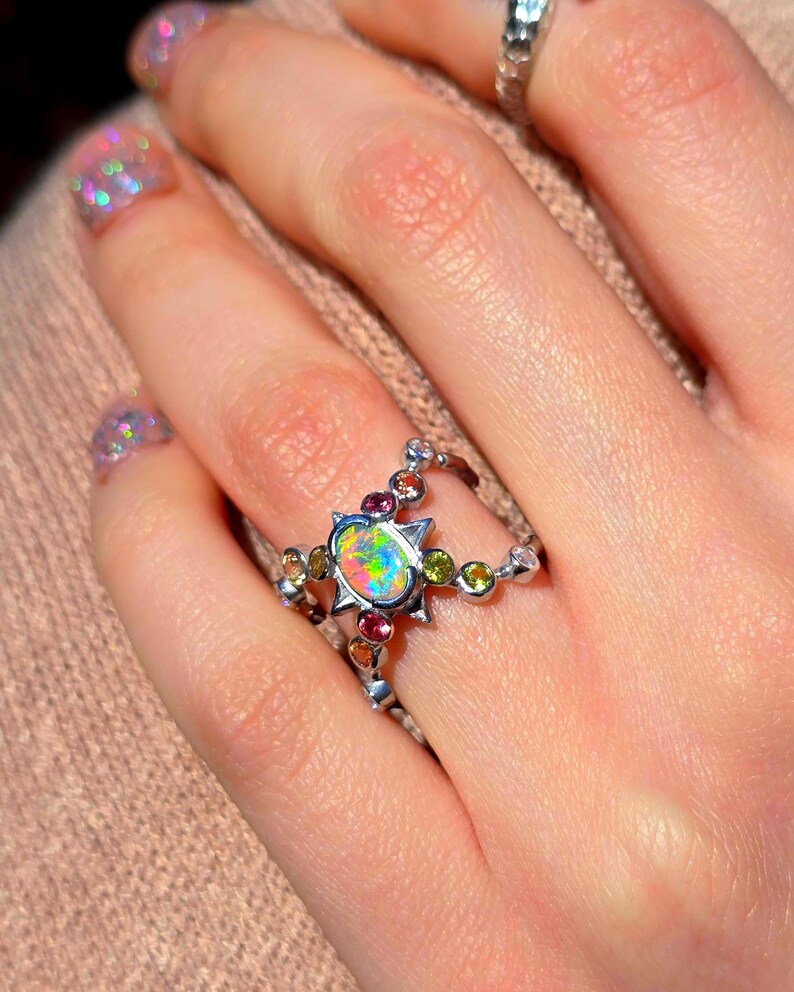 May include: A silver ring featuring a central oval opal with a vibrant play-of-colour, surrounded by a star-shaped setting. The band is accented with small, round, multi-coloured gemstones including green, pink, and orange stones.