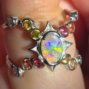May include: A silver ring featuring a central oval opal with a multi-coloured play of light, surrounded by a starburst setting. The band is accented with small, round, multi-coloured gemstones including yellow, pink, and orange stones.
