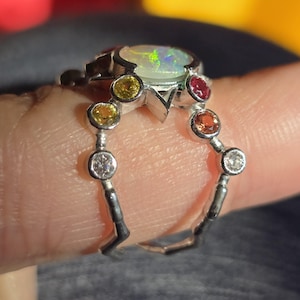 May include: A silver ring featuring a central oval opal with a play-of-colour, surrounded by a halo of small, round, multi-coloured gemstones including yellow, orange, red, and clear stones set in a pronged metal band design.