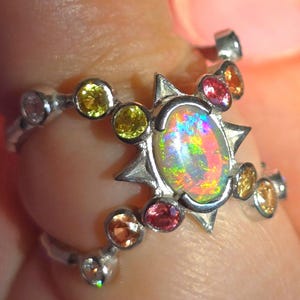 May include: A silver ring featuring a central oval-shaped opal with a vibrant play-of-colour, surrounded by a star-shaped setting. The band is accented with multiple small, round-cut gemstones in shades of yellow, orange, pink, and clear white.