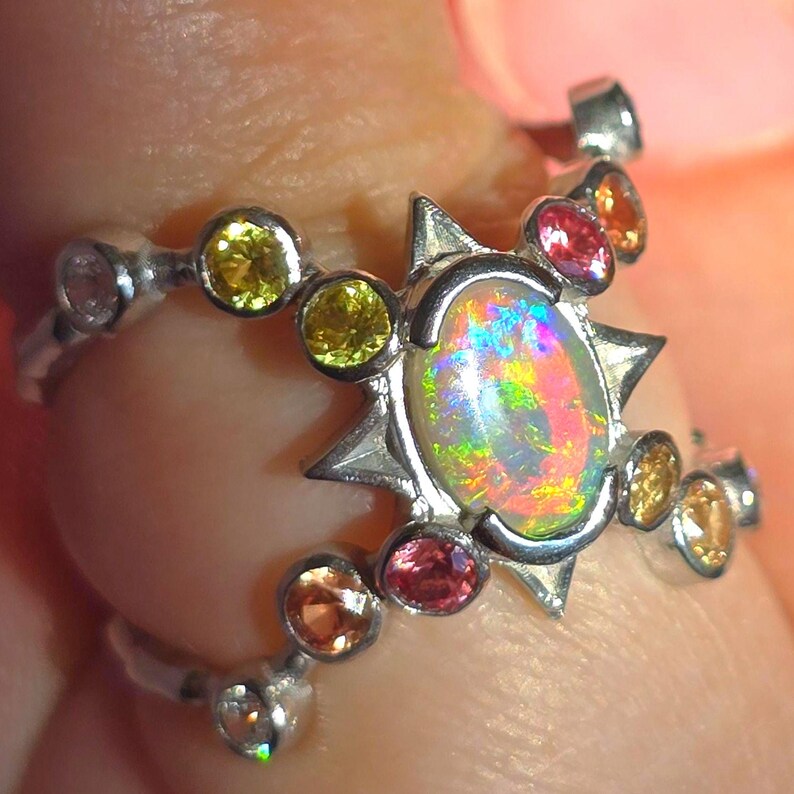 May include: A silver ring featuring a central oval-shaped opal with a vibrant play-of-colour, surrounded by a star-shaped setting. The band is accented with multiple small, round-cut gemstones in shades of yellow, orange, pink, and clear white.