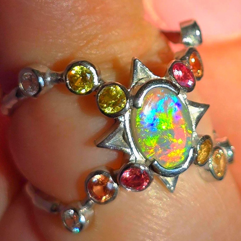 May include: A silver ring featuring a central oval opal with a vibrant play of colour, surrounded by a star-shaped setting. The band is accented with multiple small, round, multi-coloured gemstones including yellow, pink, and orange stones.