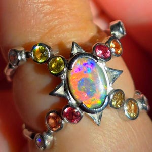 May include: A silver ring featuring a central oval-cut opal with iridescent flashes of orange, pink, and blue. The setting is accented by a starburst frame and multiple small, round-cut gemstones in shades of yellow, orange, and pink.