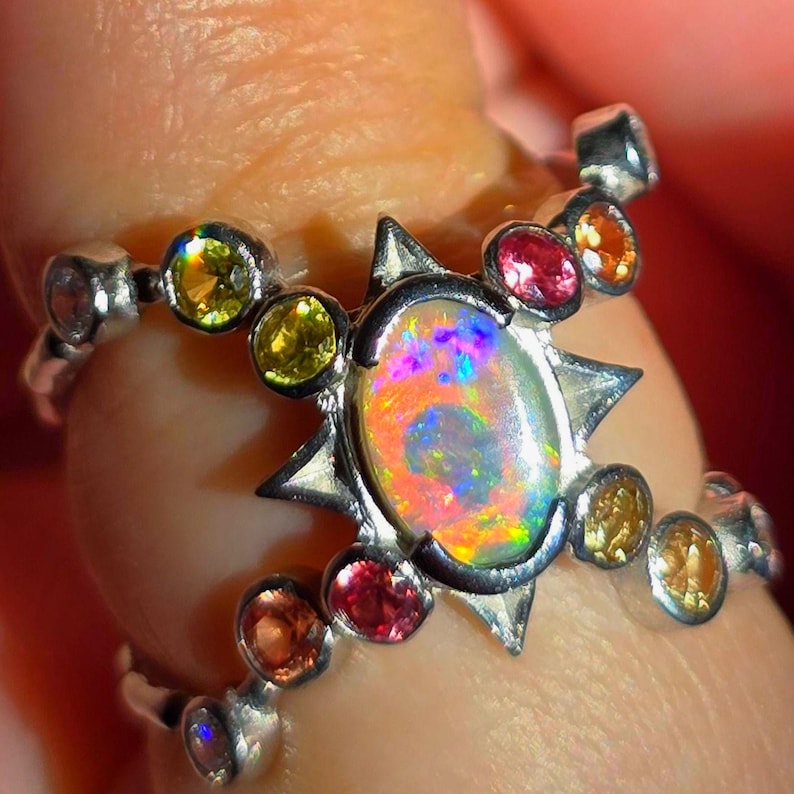 May include: A silver ring featuring a central oval-cut opal with iridescent flashes of orange, pink, and blue. The setting is accented by a starburst frame and multiple small, round-cut gemstones in shades of yellow, orange, and pink.