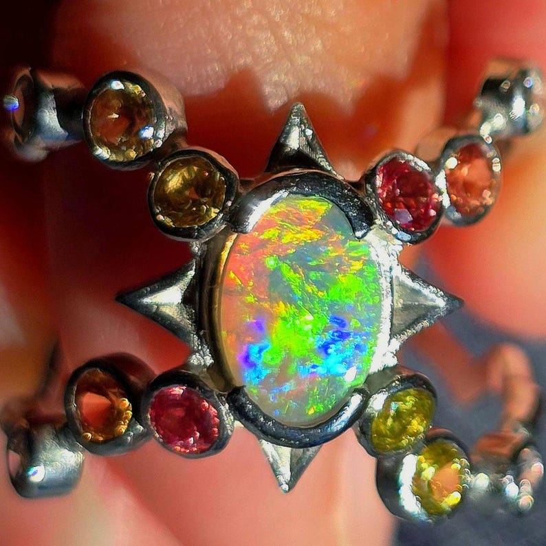 May include: A silver ring featuring a central oval opal with vibrant green, yellow, and blue flashes. The setting is accented by a star-shaped frame and multiple small, round, multi-coloured gemstones including red, orange, and yellow stones.