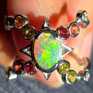 May include: A silver-toned ring featuring a central oval-shaped opal with vibrant green and orange flashes. The setting is accented by a starburst design and multiple small, round, multi-coloured gemstones including yellow, orange, and red hues.