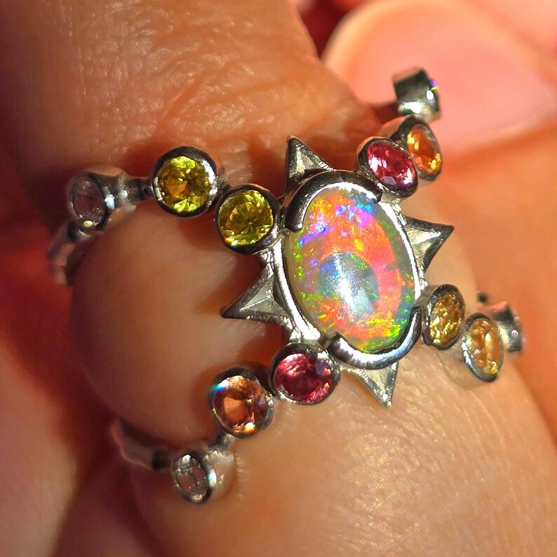 May include: A silver ring featuring a central oval opal with a play-of-colour, surrounded by a star-shaped metal setting. The band is adorned with multiple small, round, multi-coloured gemstones including yellow, orange, and pink stones.