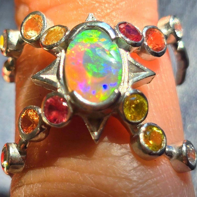 May include: A silver ring featuring a central oval-shaped opal with a vibrant play-of-colour, surrounded by a star-shaped setting. The band is accented with multiple small, round-cut gemstones in shades of orange, yellow, and red.