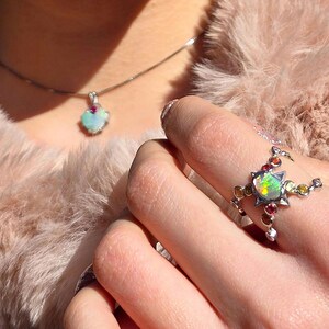 May include: A silver ring featuring a central iridescent opal surrounded by small, colourful round gemstones, paired with a matching silver necklace pendant that also displays a single opal and a small red gemstone accent.
